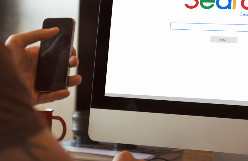 Person mit Smartphone vor Monitor mit Suchmaschine